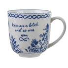 A white ceramic coffee mug with blue floral patterns and the phrase 'karma is a bitch and so are you' printed on the side, set against a plain white background.